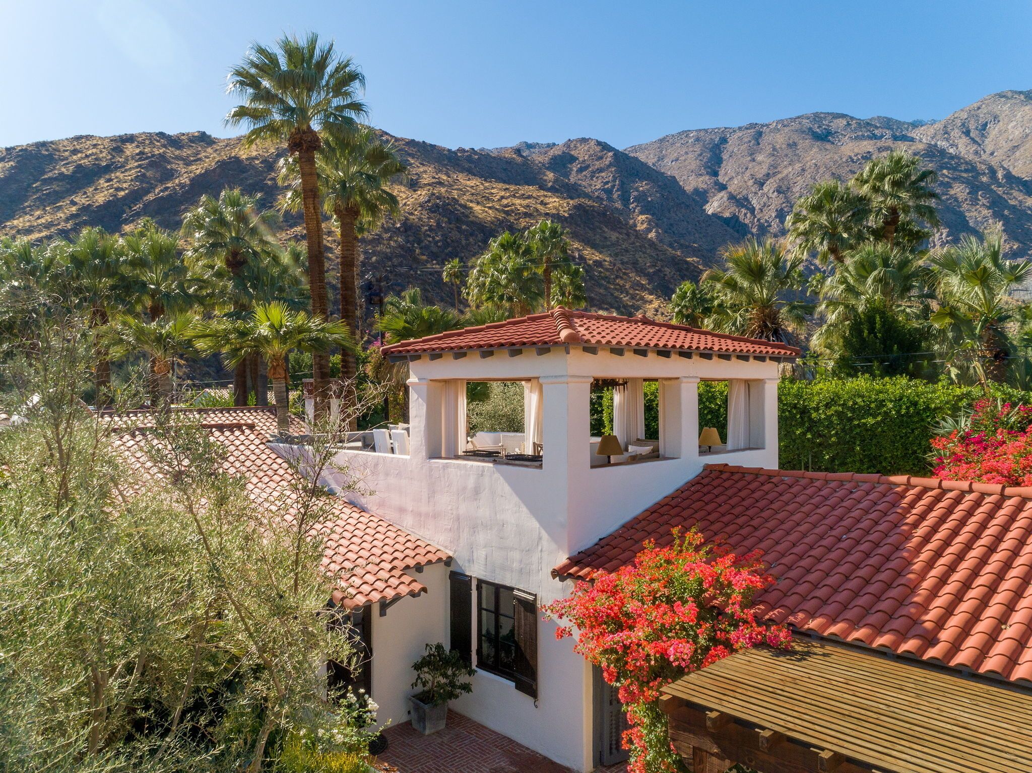 A white building with a red-tiled roof, surrounded by palm trees and mountains.