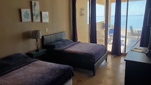 A bedroom with two beds, a nightstand with a lamp, and a view of the sea through a sliding door.