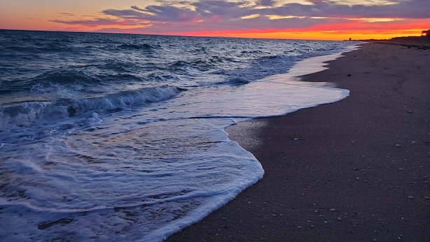 A beach with waves gently lapping onto the shore, a clear sky, and a distant horizon.