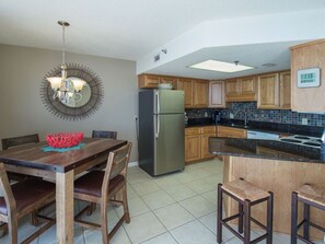 A kitchen with wooden cabinets, a stainless steel refrigerator, a dining table with chairs, and a bar area. - Beachfront 1BR/1BA | Direct Gulf Front | Resort Amenities | Sleeps 5 (Orange Beach)