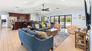 A spacious living area with a blue sofa, a coffee table, and a TV on a wooden stand. A kitchen with modern appliances is visible in the background. - Sombrero Beach | Canalfront Pool Home + Dock (Marathon)