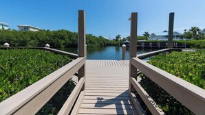 A wooden boardwalk leading to a water body, surrounded by greenery and buildings in the background. - Sombrero Beach | Canalfront Pool Home + Dock (Marathon)