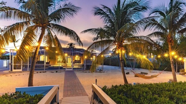 A wooden pathway leading to a beachside resort with palm trees and string lights. - Sombrero Beach | Canalfront Pool Home + Dock (Marathon)