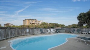 Uma piscina com espreguiçadeiras, um cercado de madeira e um edifício ao fundo.