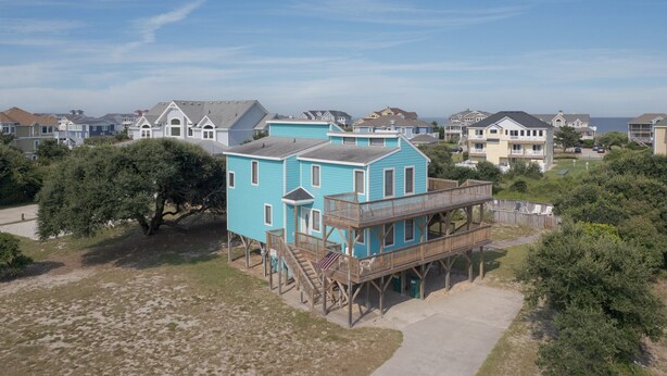 Uma área residencial à beira-mar com uma casa turquesa de destaque, um deck de madeira e uma pequena escadaria.