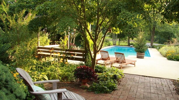 A poolside area with a wooden bench and chairs, surrounded by lush greenery and a brick pathway. - Hidden Hollow Farm - Pool; Game Rm; Sleeps 26, 45 min. to DC, Weddings/Corporate (Warrenton)
