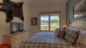 Ein Schlafzimmer mit kariertem Überwurf, einem aufgehängten Bärenfell, einem Fernseher auf einer Holzkommode und einem Fenster mit Blick auf Berge und Bäume.