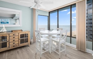 Una habitación con un ventanal que ofrece vistas al océano, un comedor blanco con sillas, un aparador de madera con cajones y un ventilador de techo.