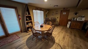 A dining area with a round wooden table and chairs, a bookshelf, and a kitchen area with a microwave and cabinets.