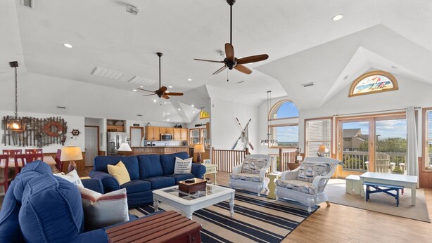 A spacious living area with a blue sofa, a white coffee table, and a wooden chair. There are ceiling fans, a kitchen in the background, and a large window with a view.