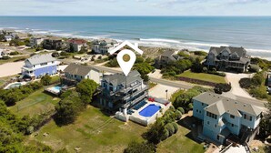 An aerial view of a coastal residential area with houses, a large swimming pool, and a beach in the background.