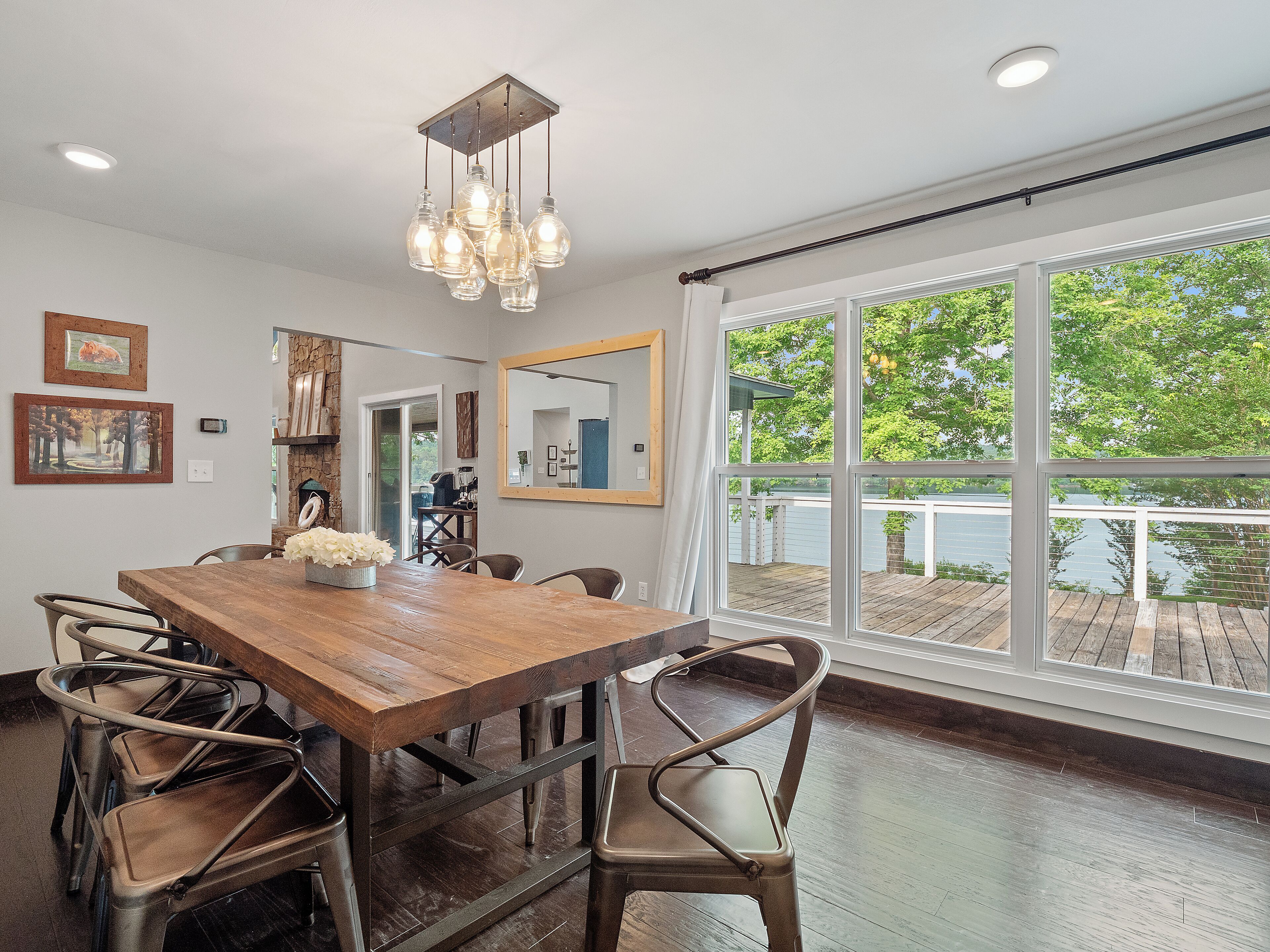 A dining area with a wooden table and metal chairs, large windows with a view of a dock and water, and artwork on the walls.