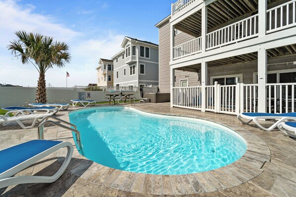 A swimming pool with lounge chairs, a palm tree, and a multi-story building with balconies. - 5409- Latitude Adjustment - Dog-Friendly Nags Head Home w/ Private Pool (Nags Head)