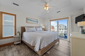 A bedroom with a large bed, a ceiling fan, a window with blinds, a small table with a lamp, and a view of houses through the window. - 5409- Latitude Adjustment - Dog-Friendly Nags Head Home w/ Private Pool (Nags Head)