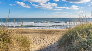 Ein Strand mit einem sandigen Pfad, der zum Meer führt, umgeben von hohem Gras und einem klaren, blauen Himmel mit vereinzelten Wolken.