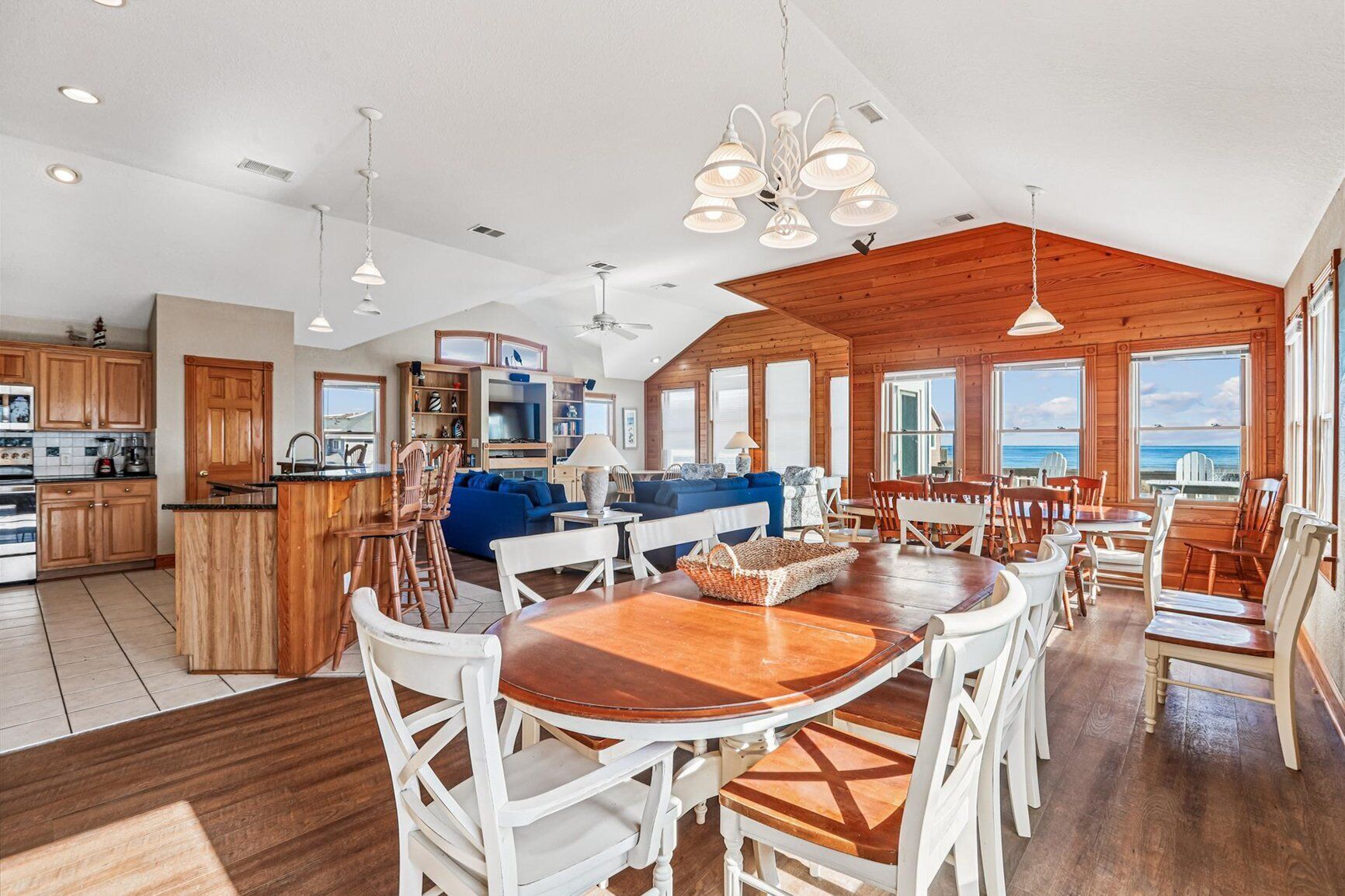 A spacious kitchen with wooden cabinets, a dining area with a large wooden table and white chairs, and a living area with a blue sofa and wooden ceiling.