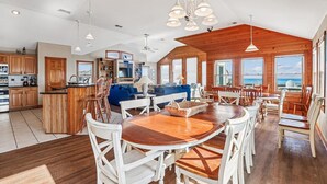 Una cocina amplia con armarios de madera, un comedor con una gran mesa de madera y sillas blancas, y una sala de estar con un sofá azul y techo de madera.