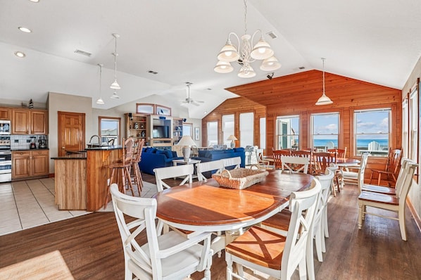 A spacious kitchen with wooden cabinets, a dining area with a large wooden table and white chairs, and a living area with a blue sofa and wooden ceiling.