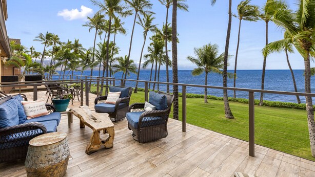 A deck with wicker furniture, a stone barrel, and a view of the ocean with palm trees.