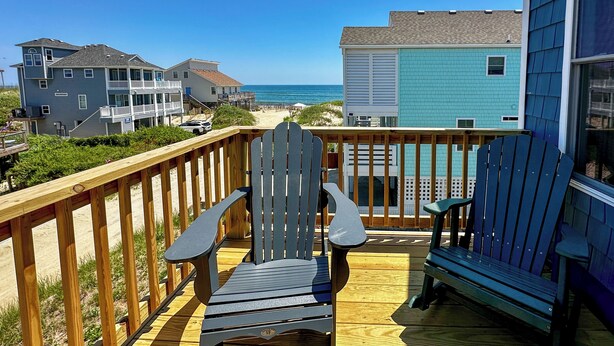 Una terraza de madera con dos sillas Adirondack y un banco, con vistas a una playa y casas.