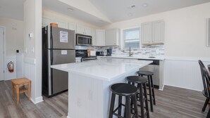Una cocina moderna con armarios blancos, electrodomésticos de acero inoxidable, una isla central con taburetes y un ventanal con persianas.