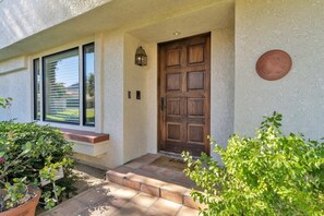 A house entrance with a wooden door, a window with blinds, a potted plant, and a small step leading to the door. - Desert Luxe | 3BR Santa Rosa Cove (La Quinta)