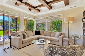 A modern living room with a beige sectional sofa, a glass coffee table, and a patterned armchair. There is a floor lamp and a small side table with a vase of flowers. Large windows provide a view of the outdoors. - Desert Luxe | 3BR Santa Rosa Cove (La Quinta)