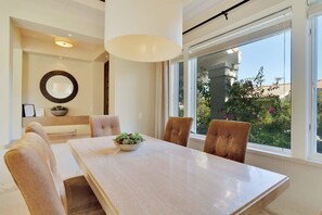 A dining area with a large table, four chairs, a bowl of flowers, and a view of greenery outside. - Desert Luxe | 3BR Santa Rosa Cove (La Quinta)