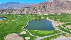 A golf course with a central pond, surrounded by green fairways and sand bunkers, with mountains in the background. - Desert Luxe | 3BR Santa Rosa Cove (La Quinta)