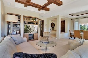 A spacious living room with a large sofa, a glass coffee table, and a patterned armchair. There is a bookshelf filled with decorative items and a dining area visible in the background. - Desert Luxe | 3BR Santa Rosa Cove (La Quinta)
