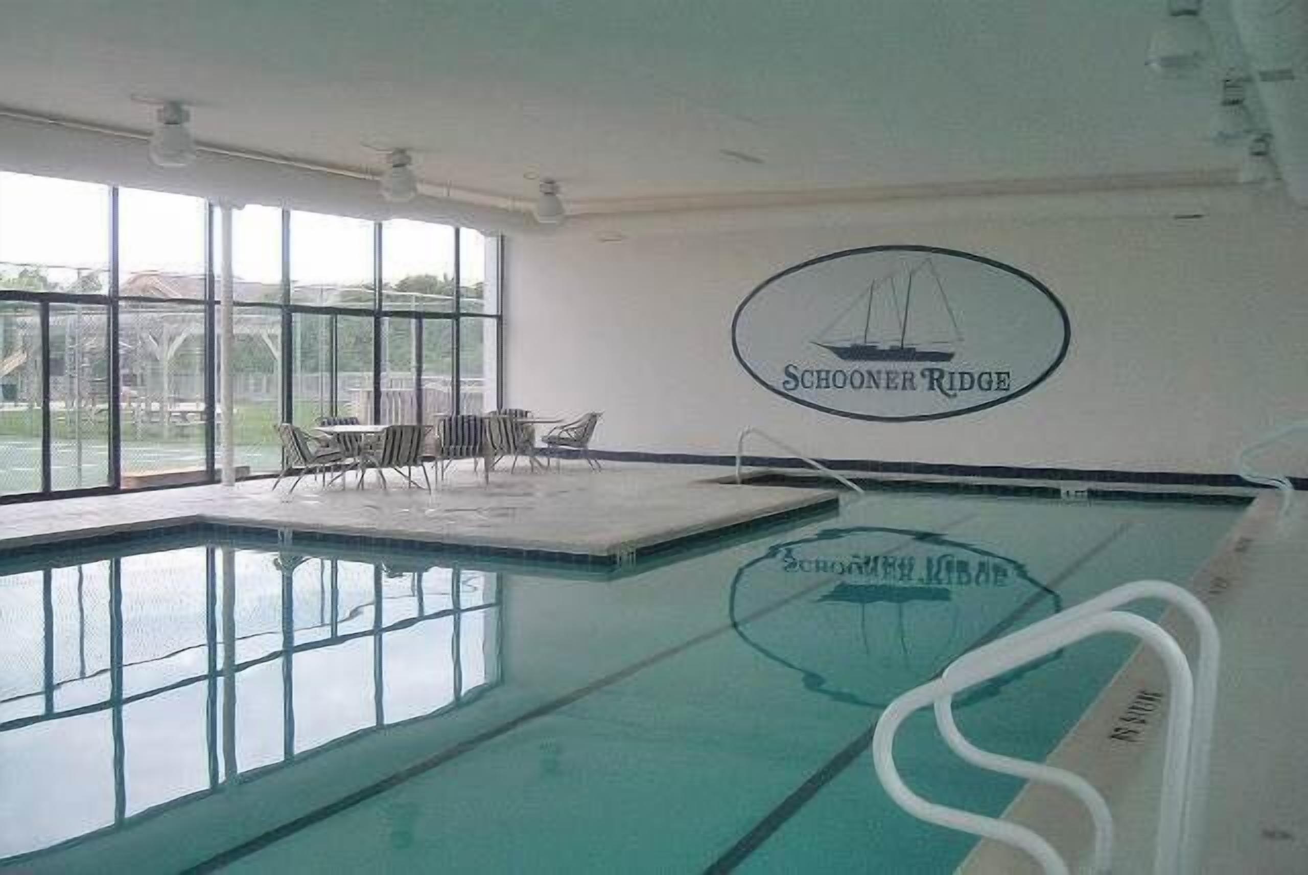 An indoor swimming pool with clear glass walls, a logo on the wall, and a seating area with chairs.