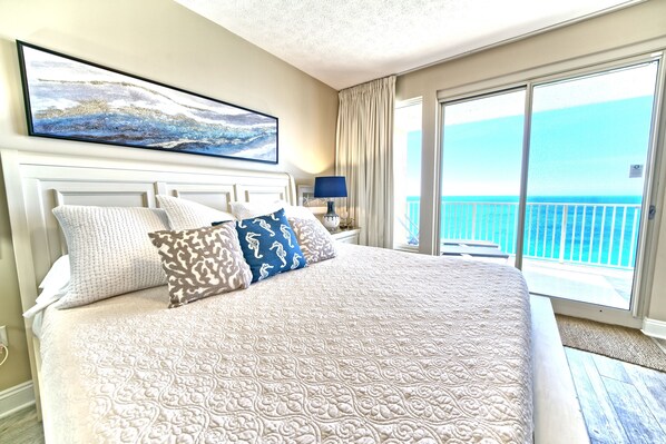Uma cama grande com colcha estampada, um quadro sobre ela e vista para o oceano através de uma varanda.