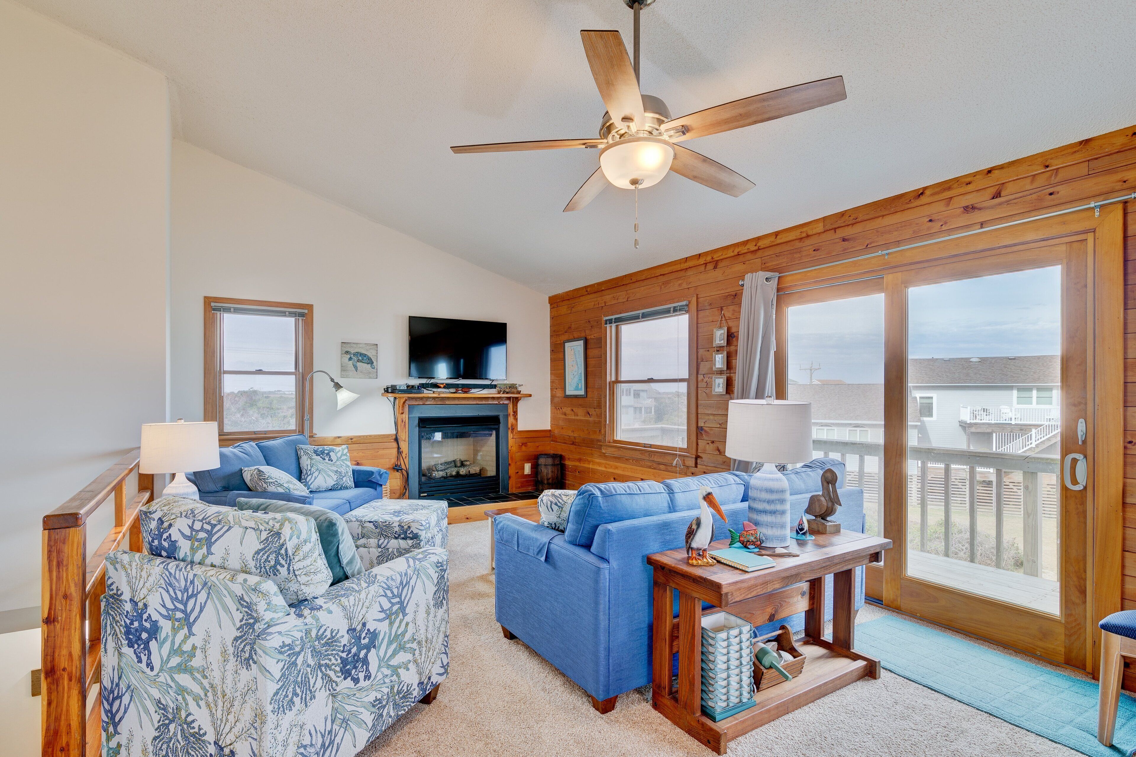 A cozy living room with a fireplace, blue sofas, a ceiling fan, and large windows overlooking a beach.