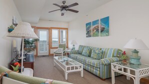 A living room with a striped sofa, a glass coffee table, a white armchair, and a ceiling fan. - C211-Mary's Heaven - Soundfront Nags Head Condo | Near Beach (Nags Head)