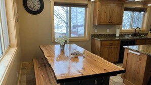 Una cocina con una mesa de madera grande, armarios de madera, un reloj y una ventana con vistas a árboles.