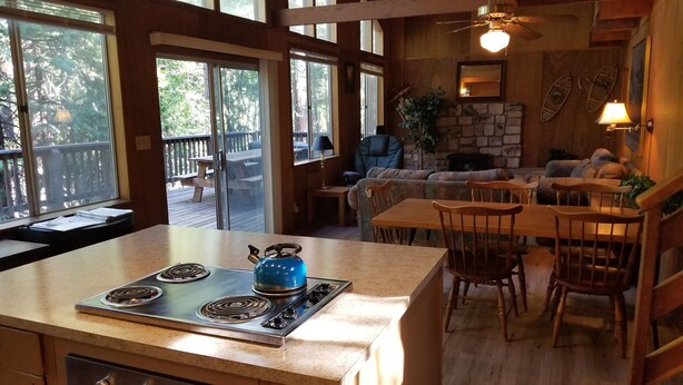 A spacious kitchen with a large island, wooden cabinets, and a blue kettle. There is a dining area with a table and chairs, and a stone fireplace. - PinecrestAreaRentals #17  Fun cabin Summer 3rd night free. Just Book 2nights. (Cold Springs)