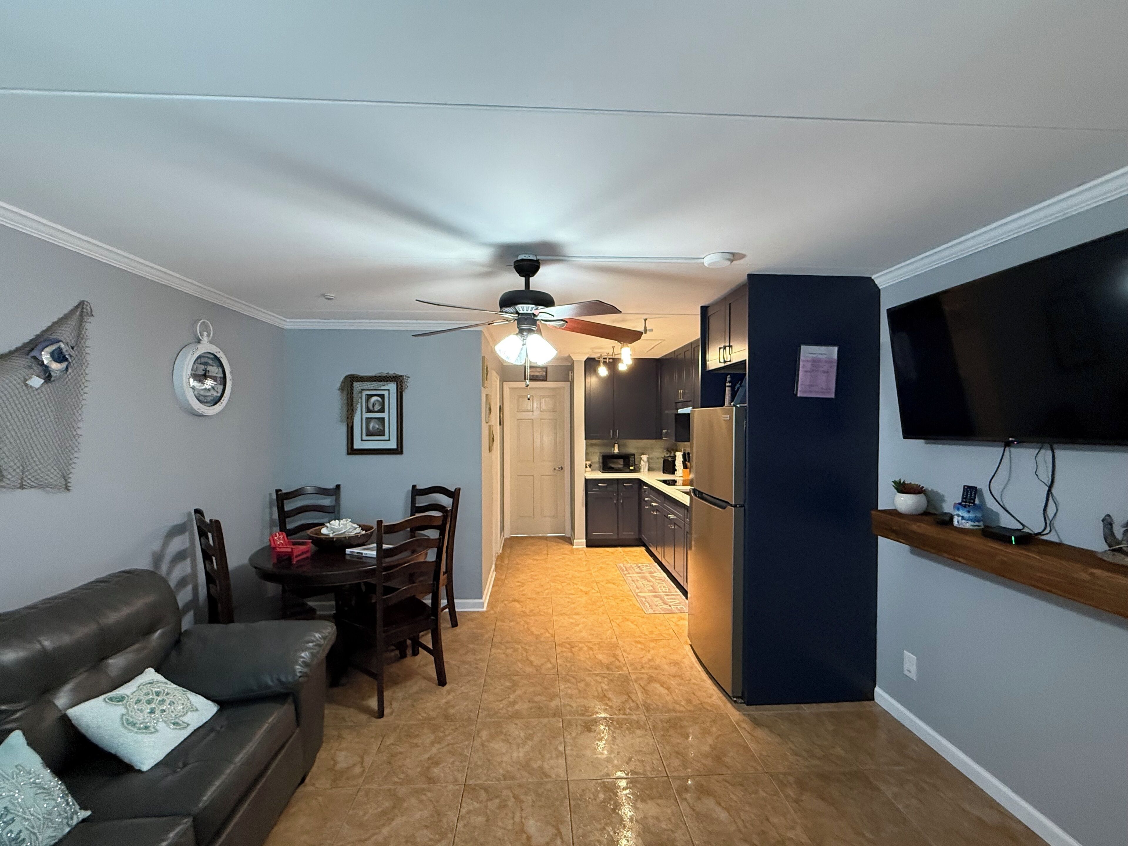 A living room with a sofa, a dining table with chairs, a kitchen area with cabinets and appliances, and a TV mounted on the wall.