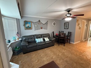 A living room with a dark leather couch, a dining table with chairs, a ceiling fan, and a window with blinds.