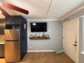 A kitchen with a stainless steel refrigerator, a flat-screen TV mounted on the wall, a wooden shelf with decorative items, and a white door.