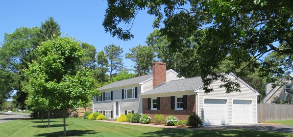 A two-story house with a garage, surrounded by a well-maintained lawn and trees. - Central A/C, Walk to Beach, Gated Yard, Wifi, HD TV, Gas Grill, Firepit (West Dennis)