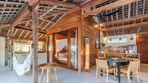 A rustic wooden interior with a hammock, a bed with a canopy, a dining area with a table and chairs, and a kitchen area.
