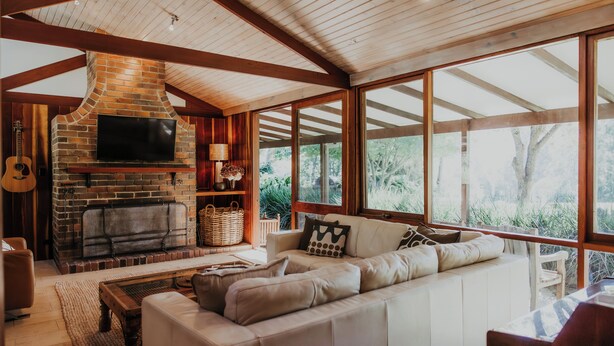 A spacious living room with a brick fireplace, a large sofa, and a television. - Blue Gum Retreat- Stunning View and Cosy (Bellingen)