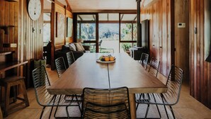 A long wooden dining table with metal chairs, surrounded by wooden walls and large windows offering a view of the outdoors. - Blue Gum Retreat- Stunning View and Cosy (Bellingen)