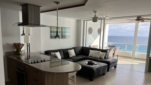 A modern kitchen with a central island, a black leather sofa, and a view of the ocean through large windows. - Ocean Front In Marbella Assigned underground Parking & fast internet 150 Mbps  (Juan Dolio)