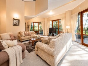 A spacious living room with a large sofa, a coffee table, and a television. There are large windows with curtains, and a balcony is visible.