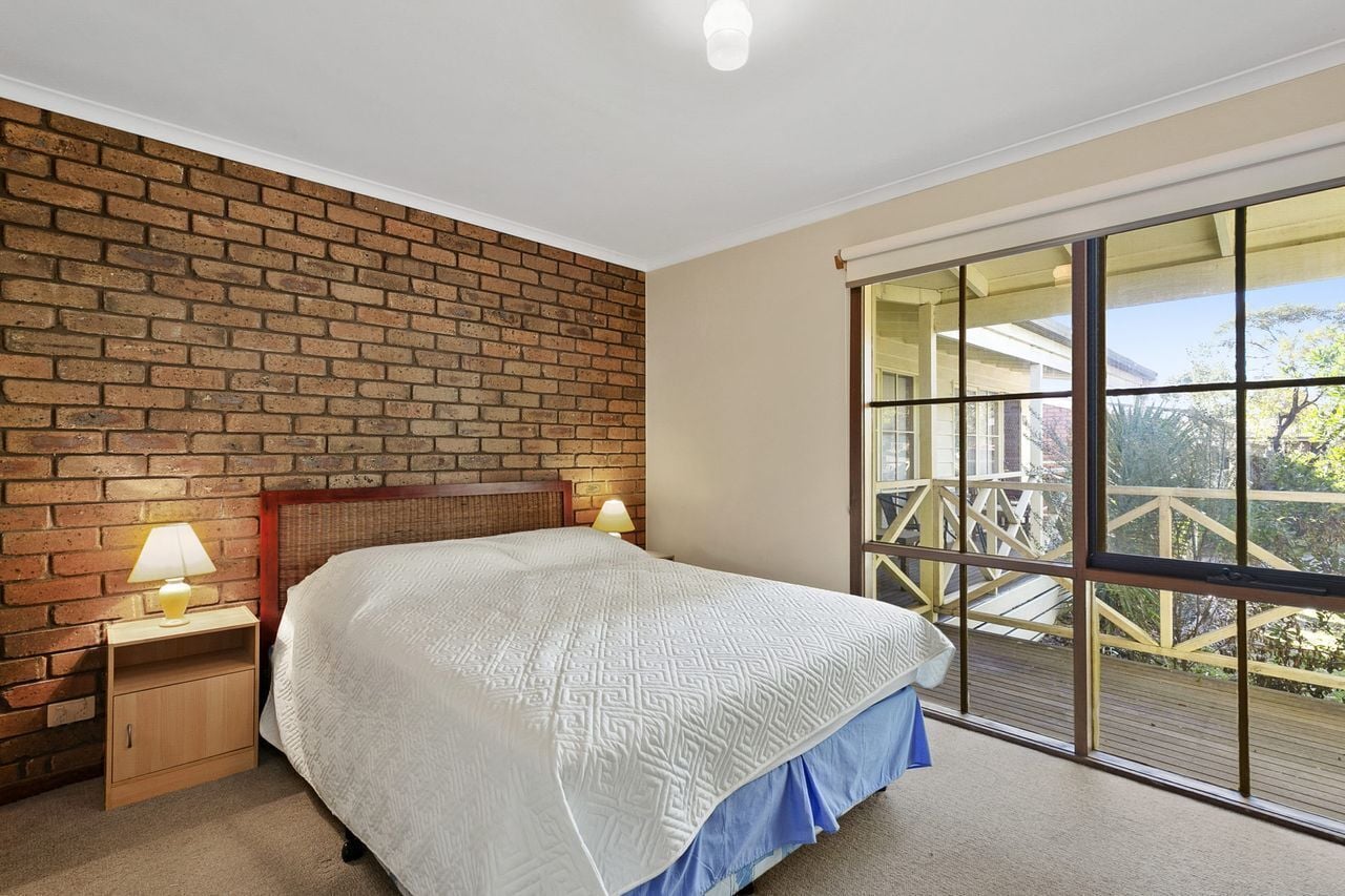 A bedroom with a brick wall, a large window with a view, a bed with a white bedspread, and a nightstand with a lamp.