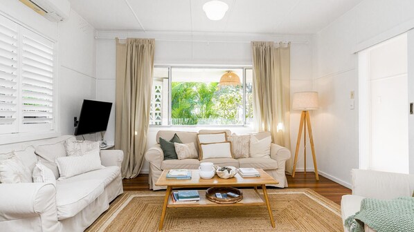 En stue med en beige sofa, et sofabord med bøger og en tekedel, et fjernsyn på væggen og et vindue med gardiner.