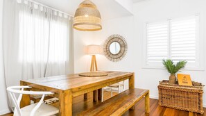 Et spiseområde med et træbord, hvide stole, en vævet pendel og gulvlampe, samt et dekorativt spejl.
