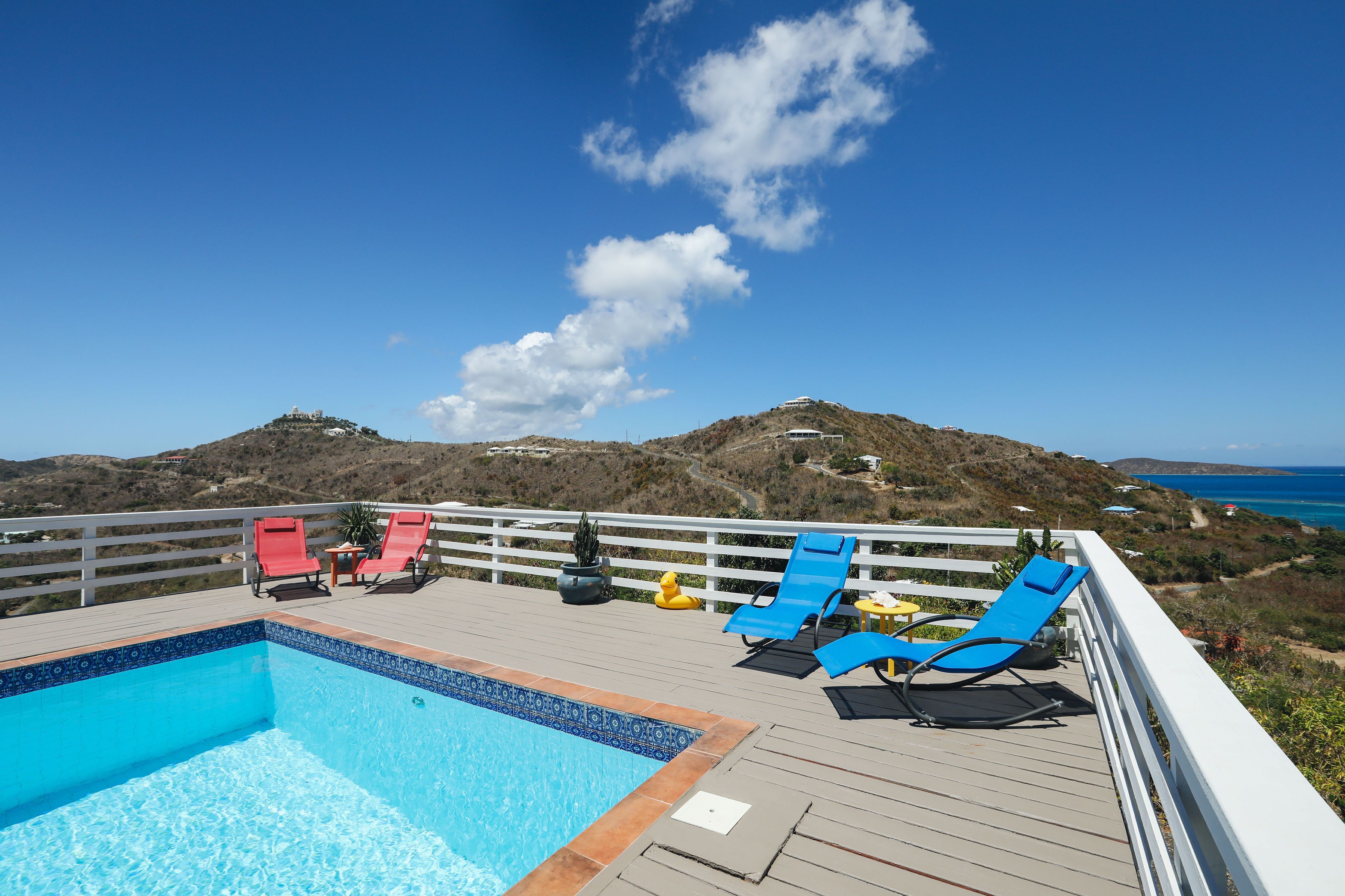 A pool area with blue and red lounge chairs, a small table, and a potted plant.