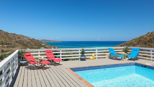A wooden deck with a pool, red and blue lounge chairs, and a view of the ocean. - Paradise Awaits (Christiansted)